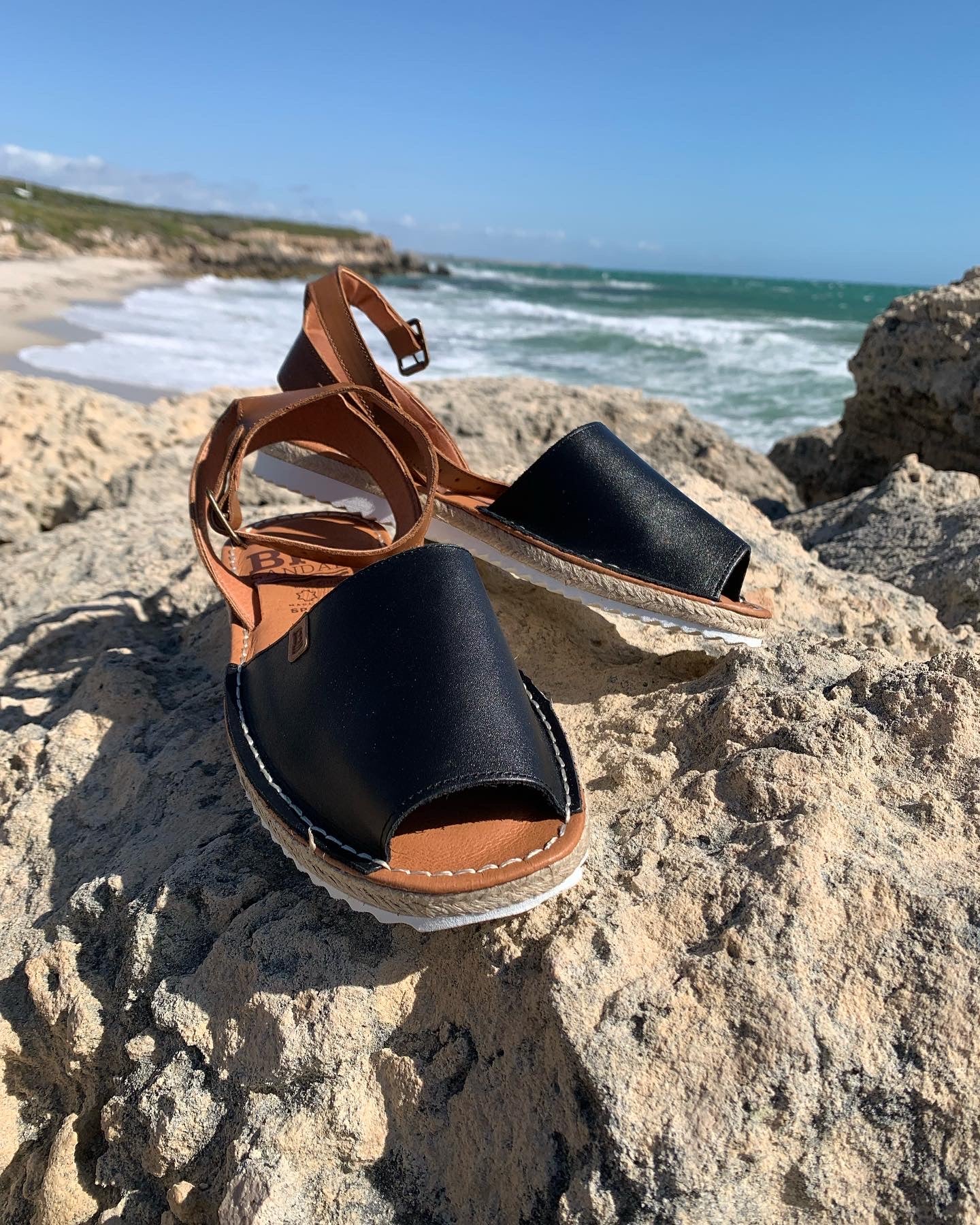 A pair of black leather sandals with tan ankle straps on a rocky beach scene , showcasing the sandals' design and fit.