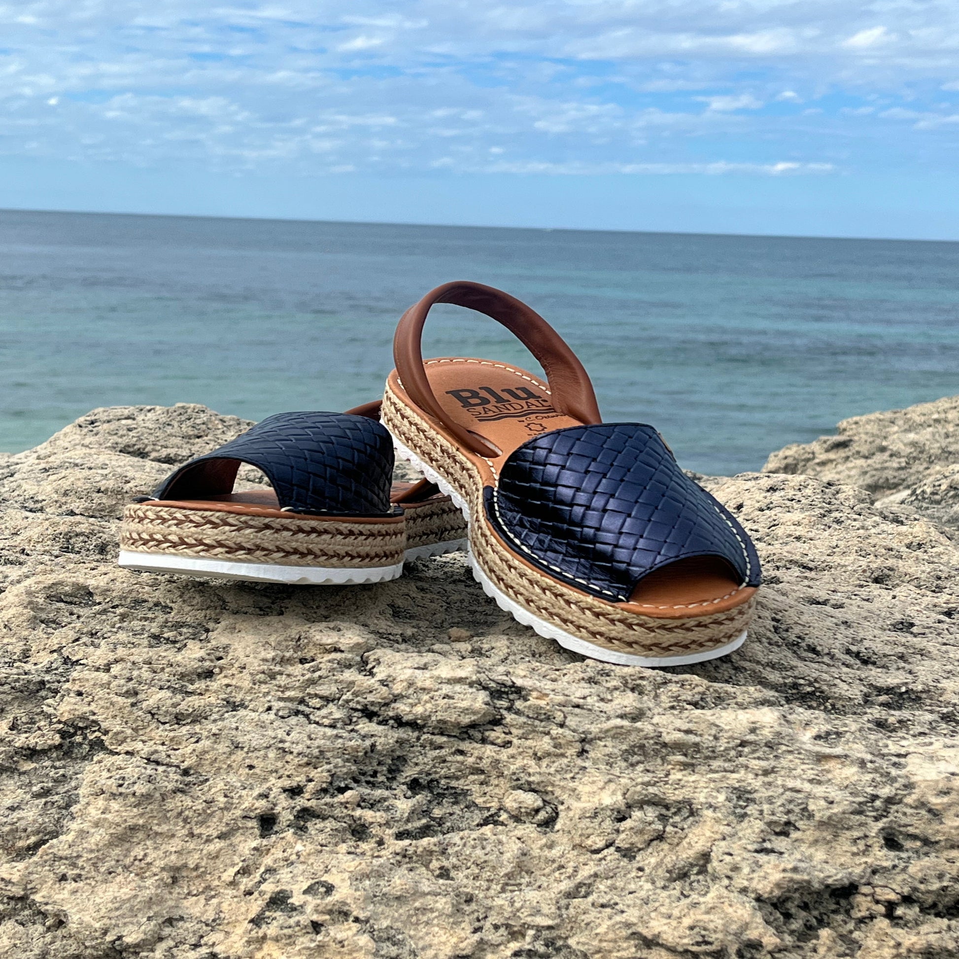 Pair of navy espadrille sandals with brown straps on a rock by the ocean.