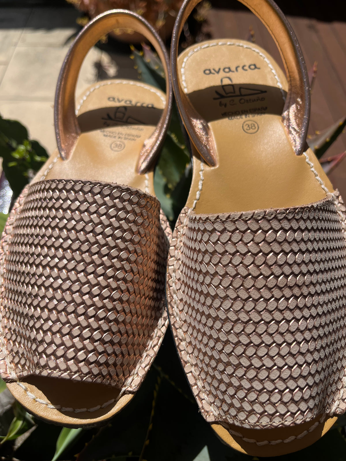 A pair of Avarca peep-toe espadrille sandals with metallic rose gold woven straps and tan leather soles, shown outdoors on a stone surface with green foliage in the background.