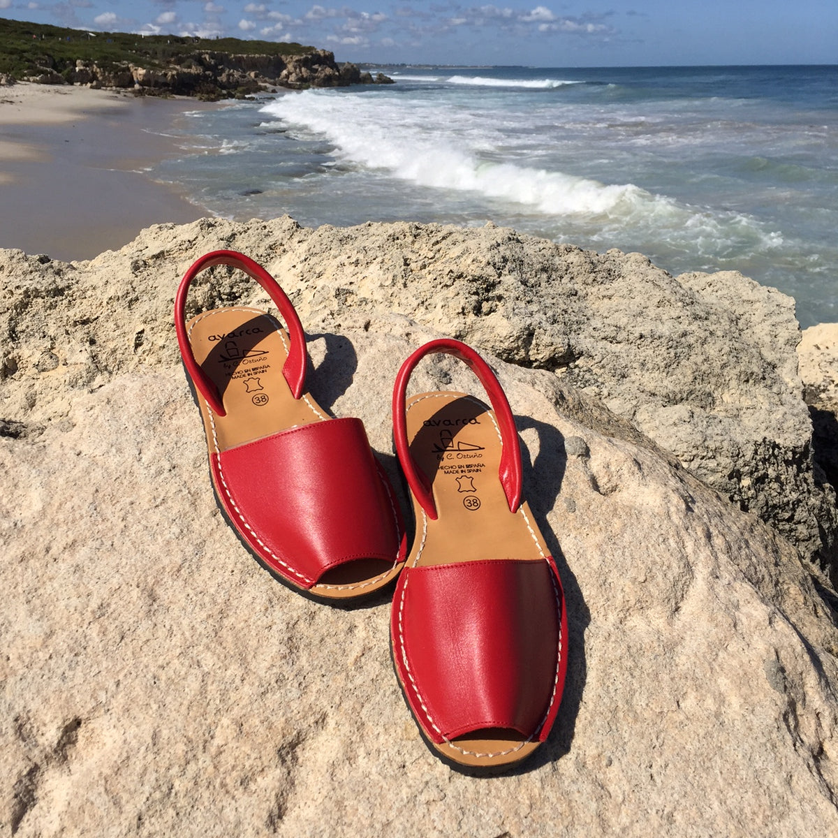 A pair of red leather ladies' sandals placed on a rocky beach with the ocean in the background.